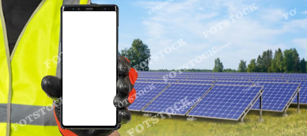 На переднем плане изображён смартфон с пустым экраном, который держит человек в защитной жилетке и перчатке. На заднем плане видны солнечные панели на фоне зелёного поля и голубого неба. Снимок иллюстрирует соединение современных технологий и возобновляемой энергетики.